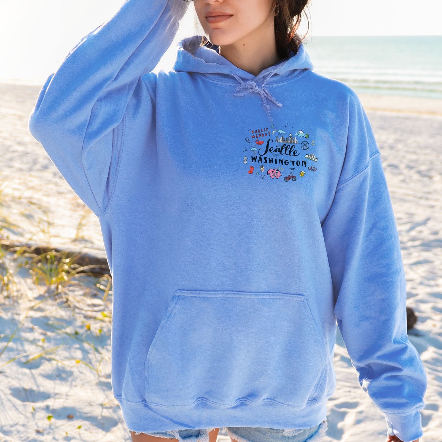 A person standing on a sandy beach wearing a light blue pullover hoodie with a small multicolor graphic on the left chest that reads 'Seattle Washington'.