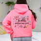 A woman viewed from behind wearing a bright pink hoodie with a colorful 'Seattle Washington' graphic on the back, paired with light blue jeans in a cozy bedroom setting.