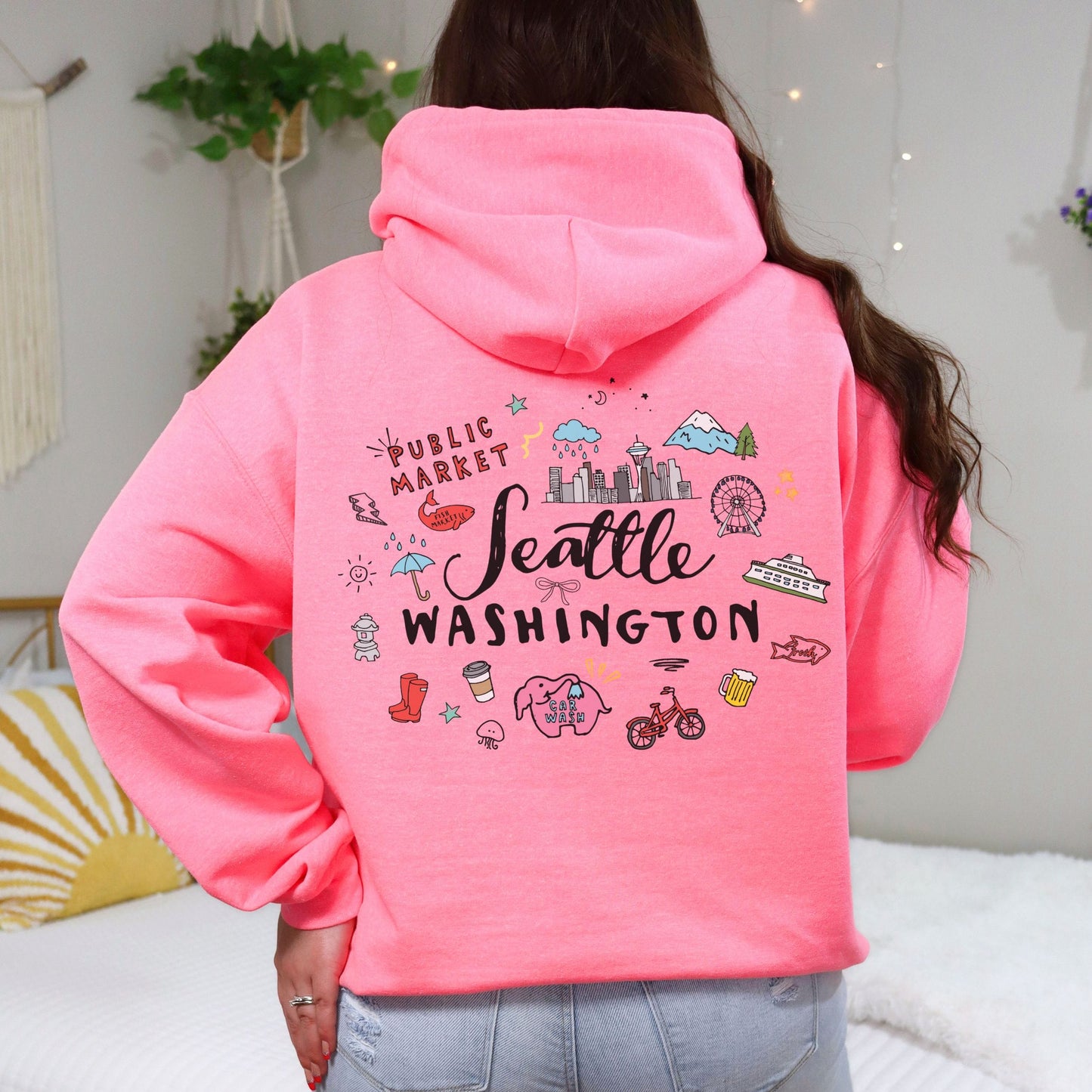 A woman viewed from behind wearing a bright pink hoodie with a colorful 'Seattle Washington' graphic on the back, paired with light blue jeans in a cozy bedroom setting.