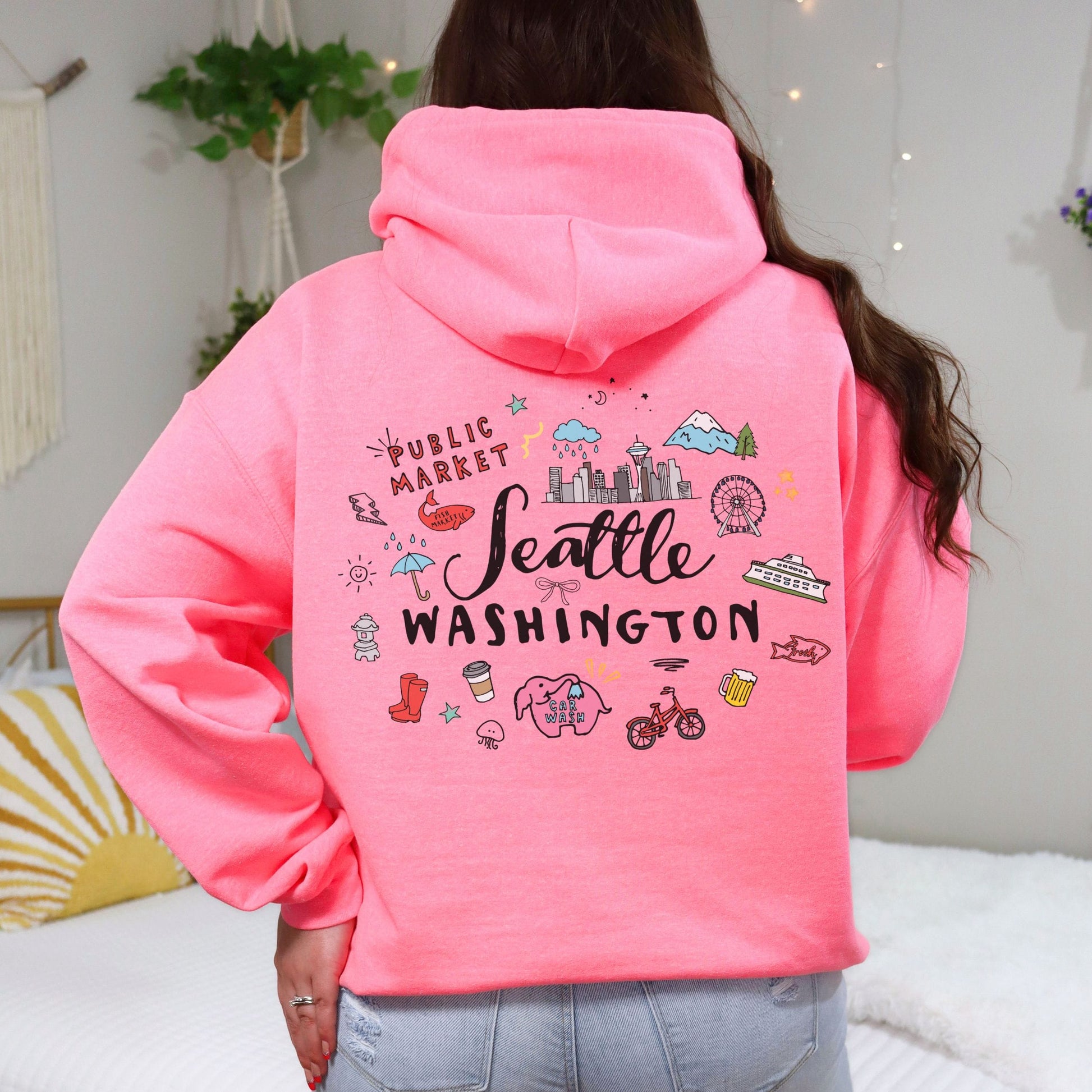 A woman viewed from behind wearing a bright pink hoodie with a colorful 'Seattle Washington' graphic on the back, paired with light blue jeans in a cozy bedroom setting.