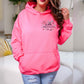 A woman wearing a bright pink pullover hoodie with a 'Seattle Washington' graphic on the chest, paired with distressed light blue jeans, standing in a brightly lit indoor setting.