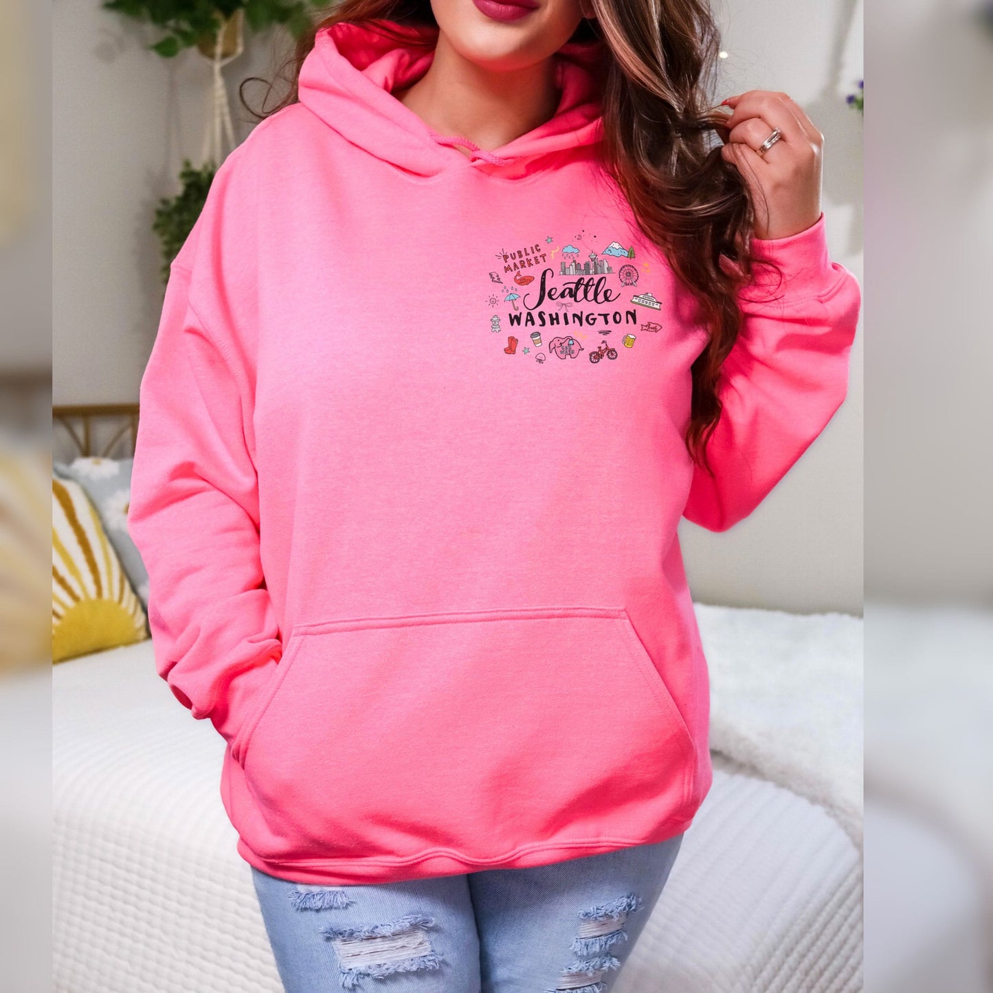 A woman wearing a bright pink pullover hoodie with a 'Seattle Washington' graphic on the chest, paired with distressed light blue jeans, standing in a brightly lit indoor setting.