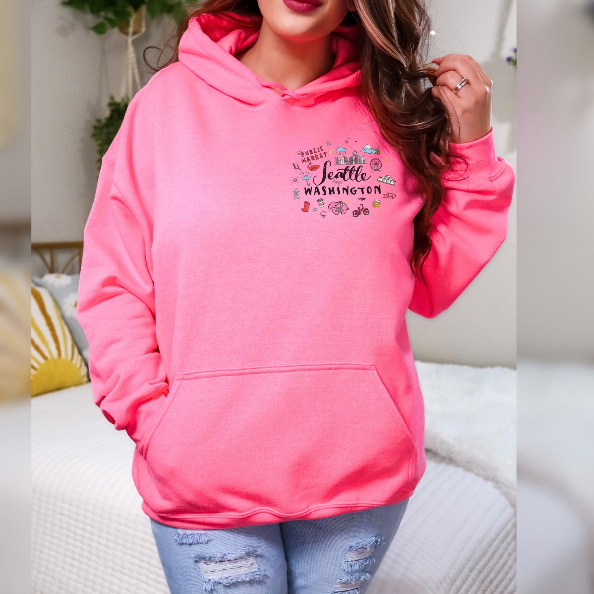 A woman wearing a bright pink pullover hoodie with a 'Seattle Washington' graphic on the chest, paired with distressed light blue jeans, standing in a brightly lit indoor setting.