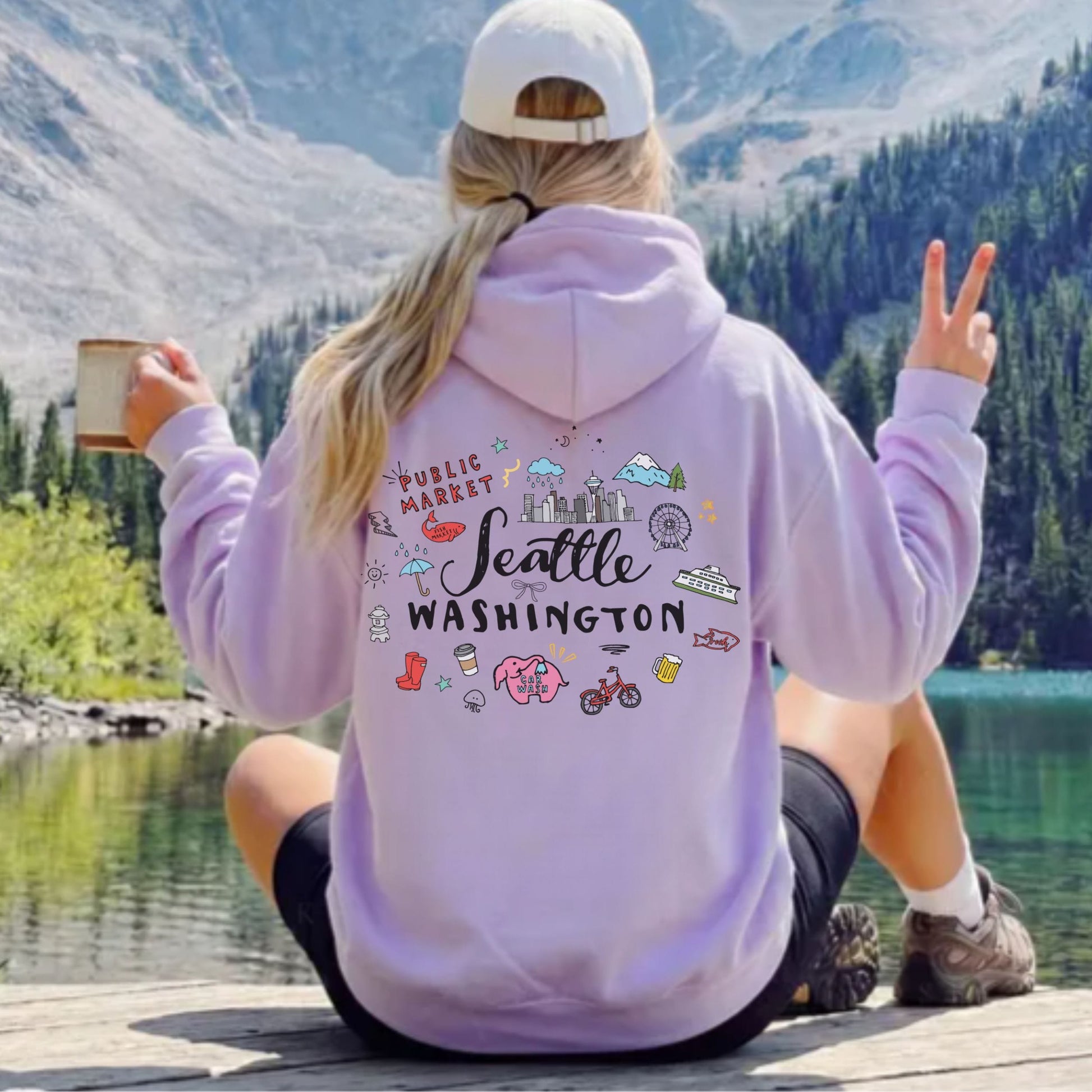 A woman sitting on a wooden dock by a mountain lake, seen from behind, wearing a light purple hoodie with a colorful 'Seattle Washington' graphic on the back, a white cap, and black shorts.
