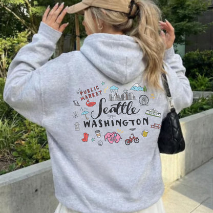 A woman viewed from behind wearing a light heather gray hoodie with a colorful back graphic that reads 'Seattle Washington' and includes small illustrated icons. She is outdoors near greenery and a concrete planter, holding a tan cap and carrying a black shoulder bag.