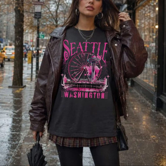 A woman stands on a wet city sidewalk wearing a dark graphic T-shirt with pink 'Seattle Washington' lettering and a Ferris wheel design, layered under a brown leather jacket. The background shows a rainy urban street with trees, storefronts, and yellow taxis.