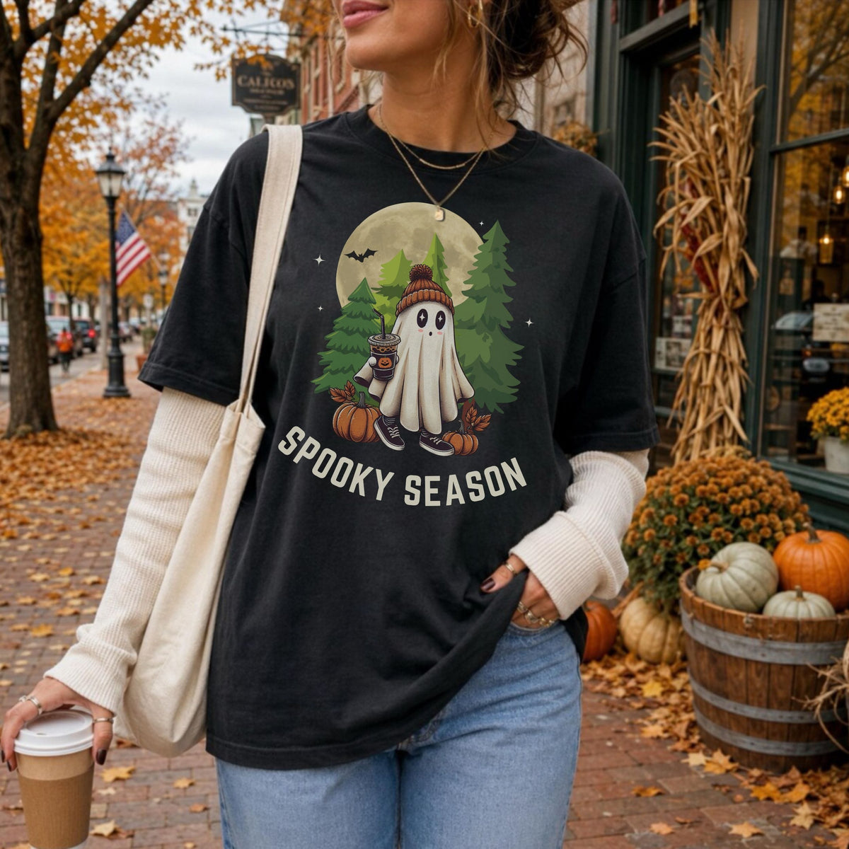 A woman wearing a black graphic t-shirt with a 'Spooky Season' cartoon ghost design, styled with a cream ribbed long-sleeve shirt underneath and blue jeans, standing on an autumn street.