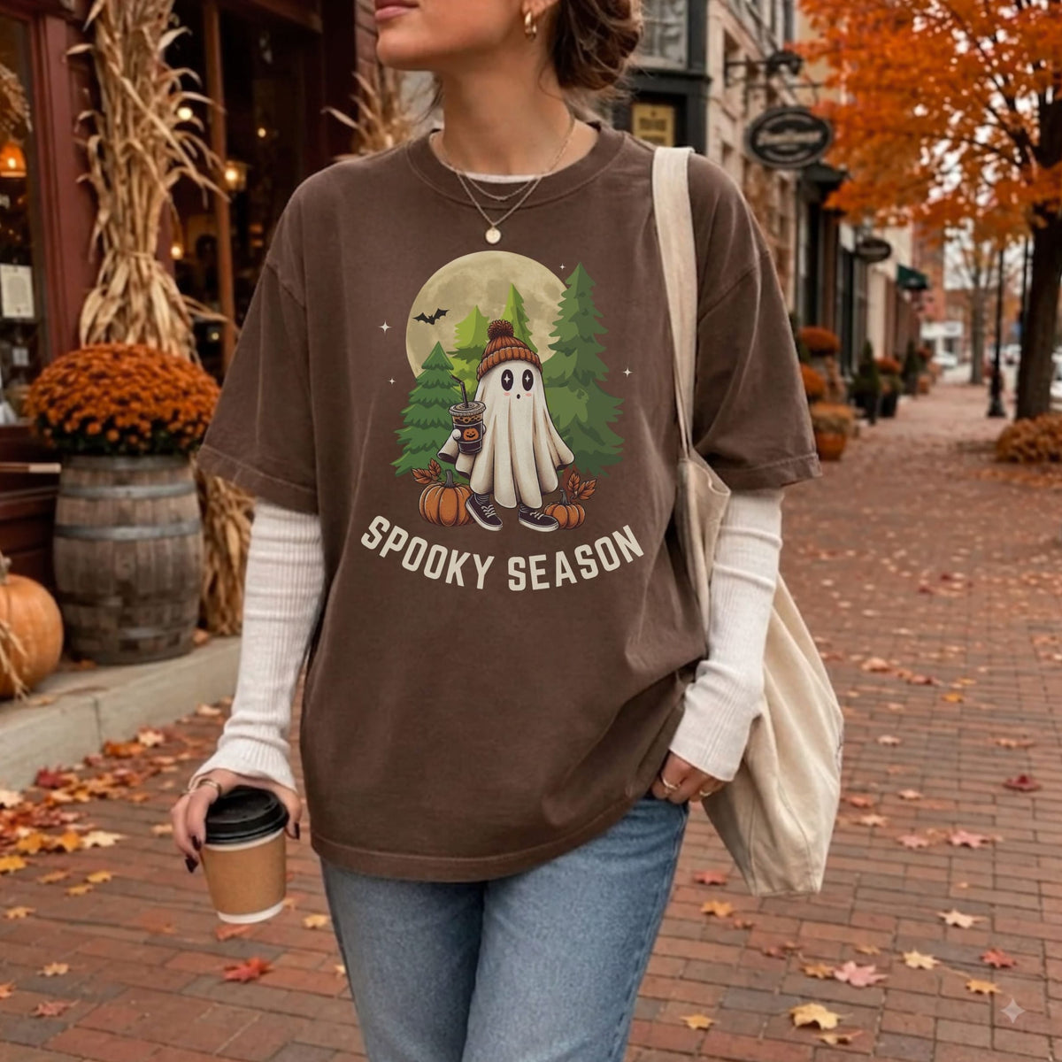 A woman wearing an oversized brown graphic T-shirt with a Halloween ghost design and the words 'SPOOKY SEASON,' layered over a white long-sleeve shirt, standing on a brick sidewalk with autumn leaves and fall decorations in the background.