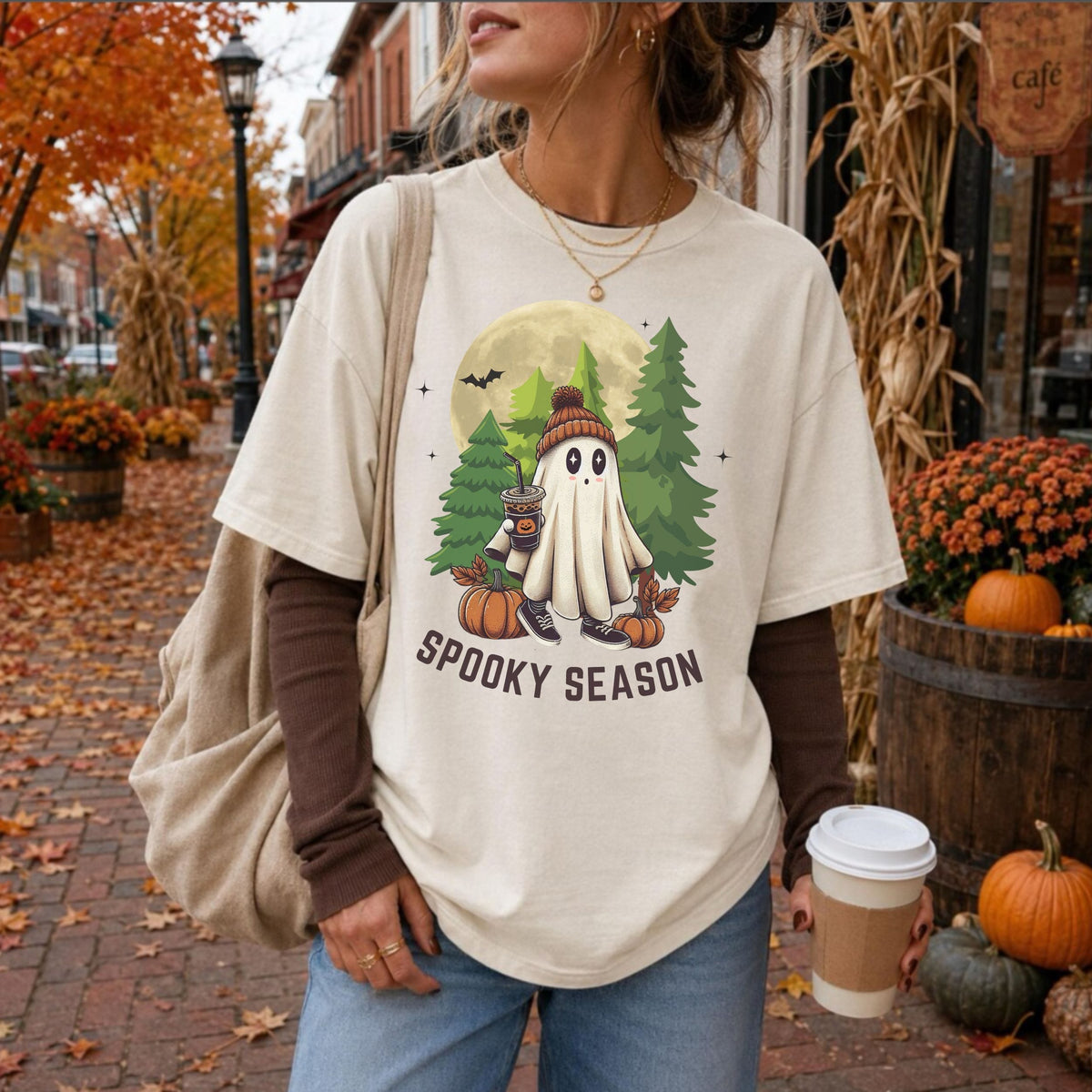 A woman wearing a cream-colored graphic t-shirt with a 'Spooky Season' Halloween ghost design, layered over a brown long-sleeve shirt, standing on a brick sidewalk in an autumn street setting.
