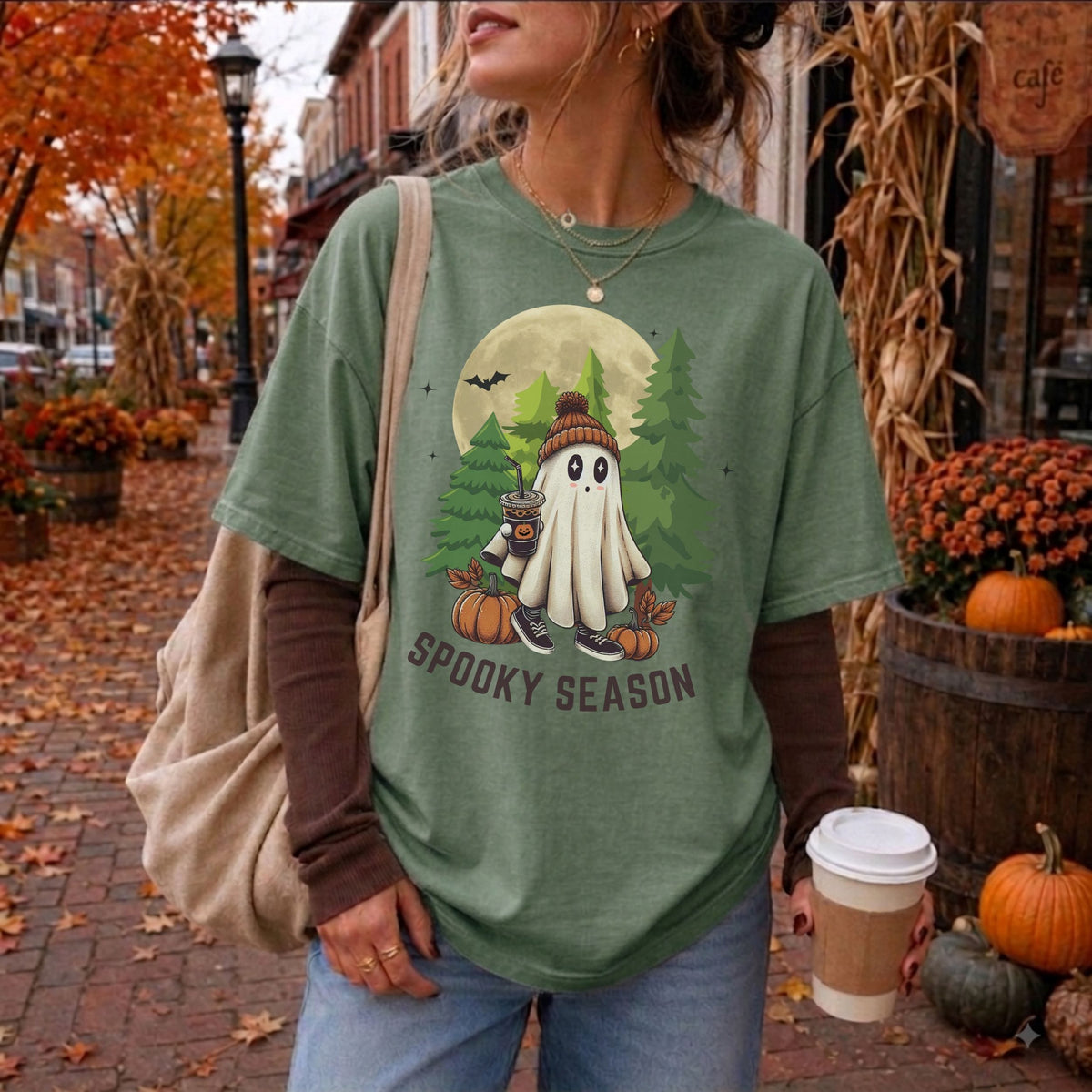 A woman wearing a moss green graphic t-shirt with a 'Spooky Season' cartoon ghost design, styled with a brown long-sleeve shirt, blue jeans, and a beige tote bag in an autumn street setting.