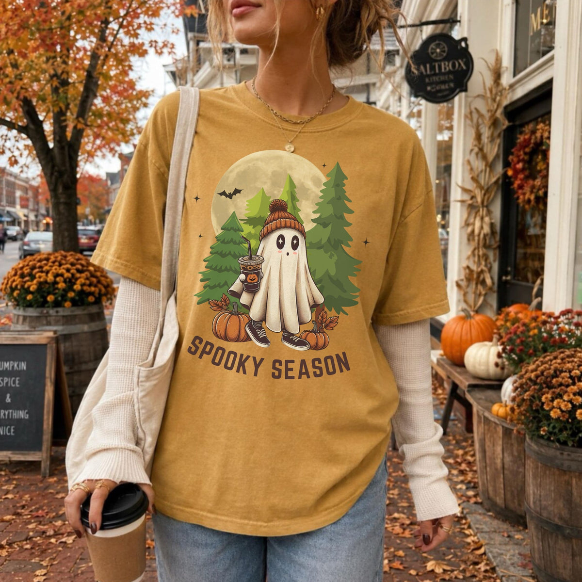 A woman wearing a mustard yellow graphic t-shirt with a 'SPOOKY SEASON' cartoon ghost design, layered over a cream ribbed long-sleeve shirt, standing on a fall-themed street sidewalk.