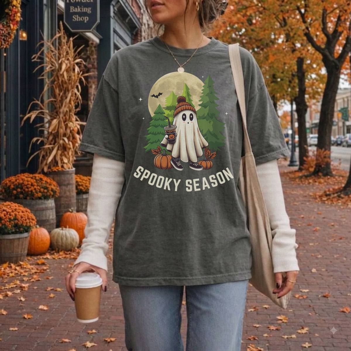 A woman wearing a faded gray graphic t-shirt with a 'Spooky Season' Halloween ghost design, layered over a brown long-sleeve shirt, standing on a brick sidewalk in an autumn street setting.