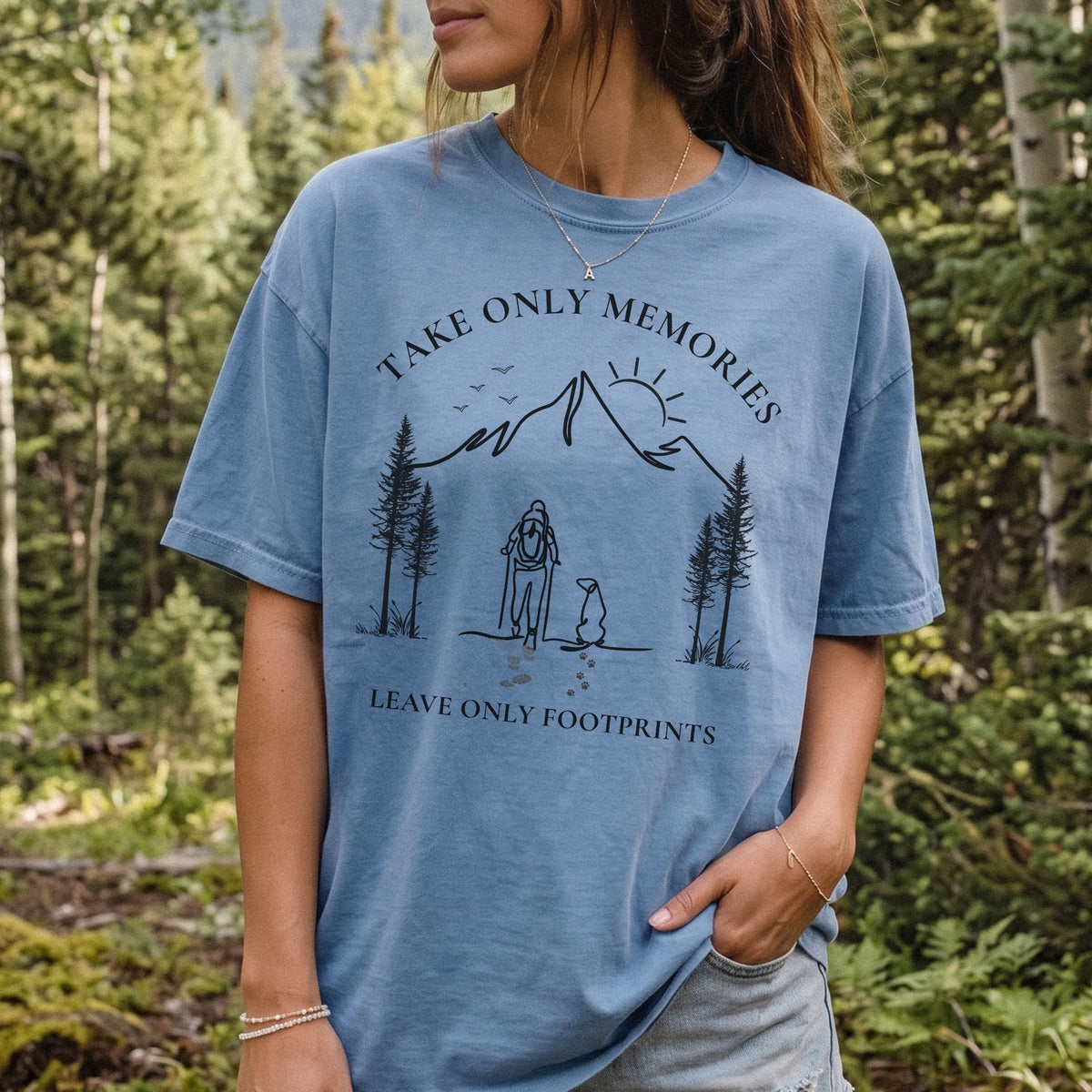 A woman wearing a faded blue graphic t-shirt with a black line art design of a mountain, trees, and a person sitting by a campfire, with the text 'TAKE ONLY MEMORIES' and 'LEAVE ONLY FOOTPRINTS'. The background is a blurred outdoor forest scene.