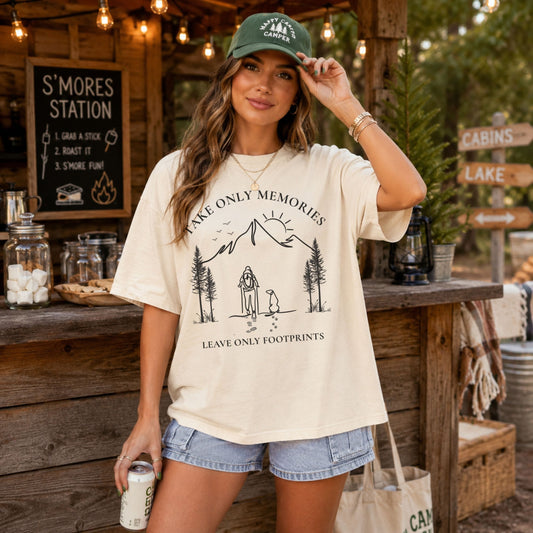 A woman stands at an outdoor rustic campsite or s'mores station wearing a cream graphic t-shirt with a black mountain and campfire design that reads 'TAKE ONLY MEMORIES' and 'LEAVE ONLY FOOTPRINTS'. She is also wearing a green 'S'mores Camp' cap and denim shorts.