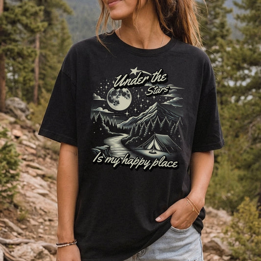 Woman wearing a black Under the Stars camping T-shirt with a moonlit mountain, forest, and night sky graphic.