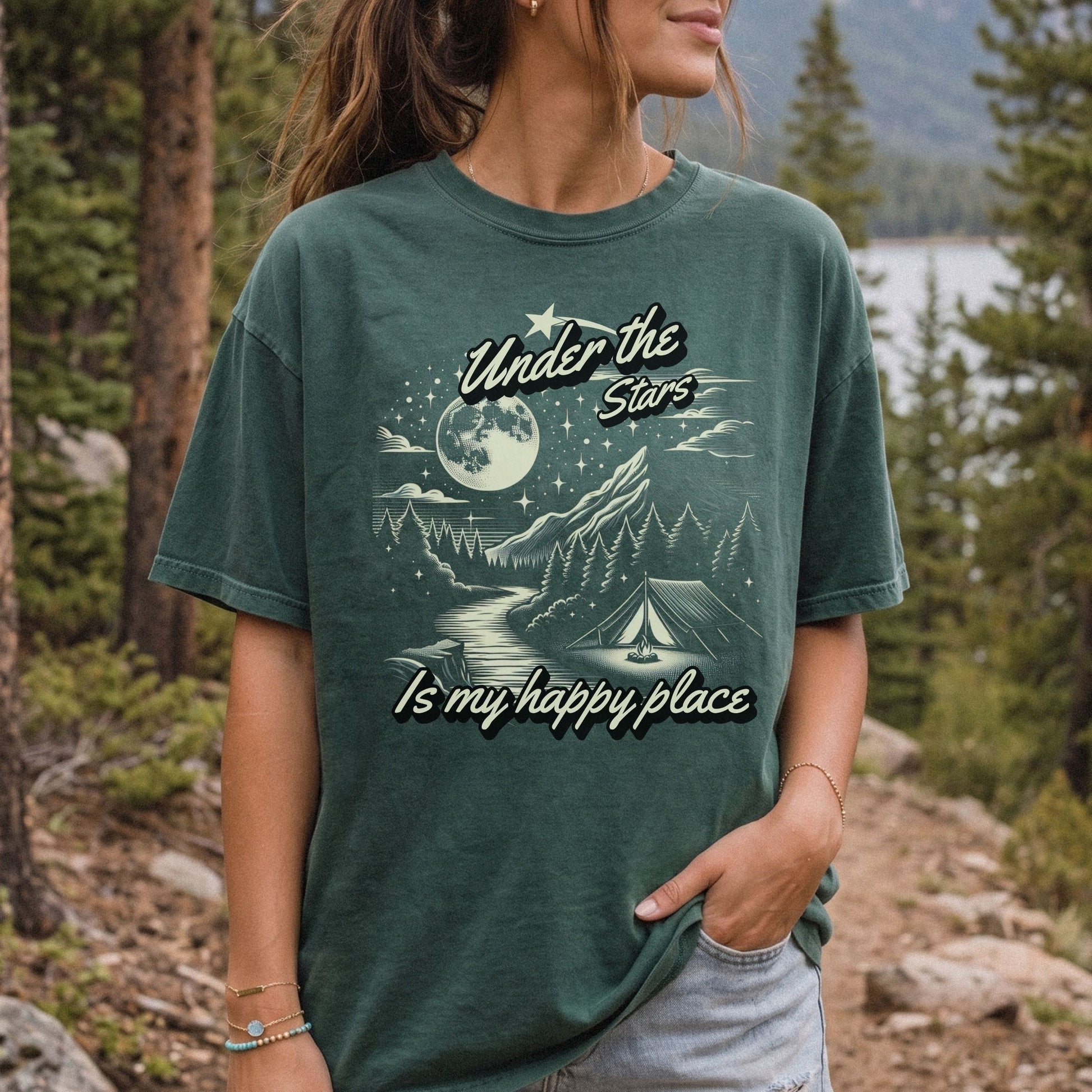 A woman wearing a dark green graphic t-shirt with a camping moonlit scene and the text 'Under the Stars Is my happy place', photographed outdoors in a forest setting.