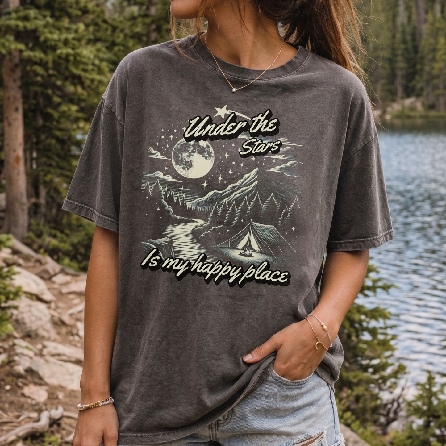 A woman wearing a dark gray mineral-wash graphic t-shirt with a camping scene and the text 'Under the Stars Is my happy place', standing outdoors near a lake and pine trees.