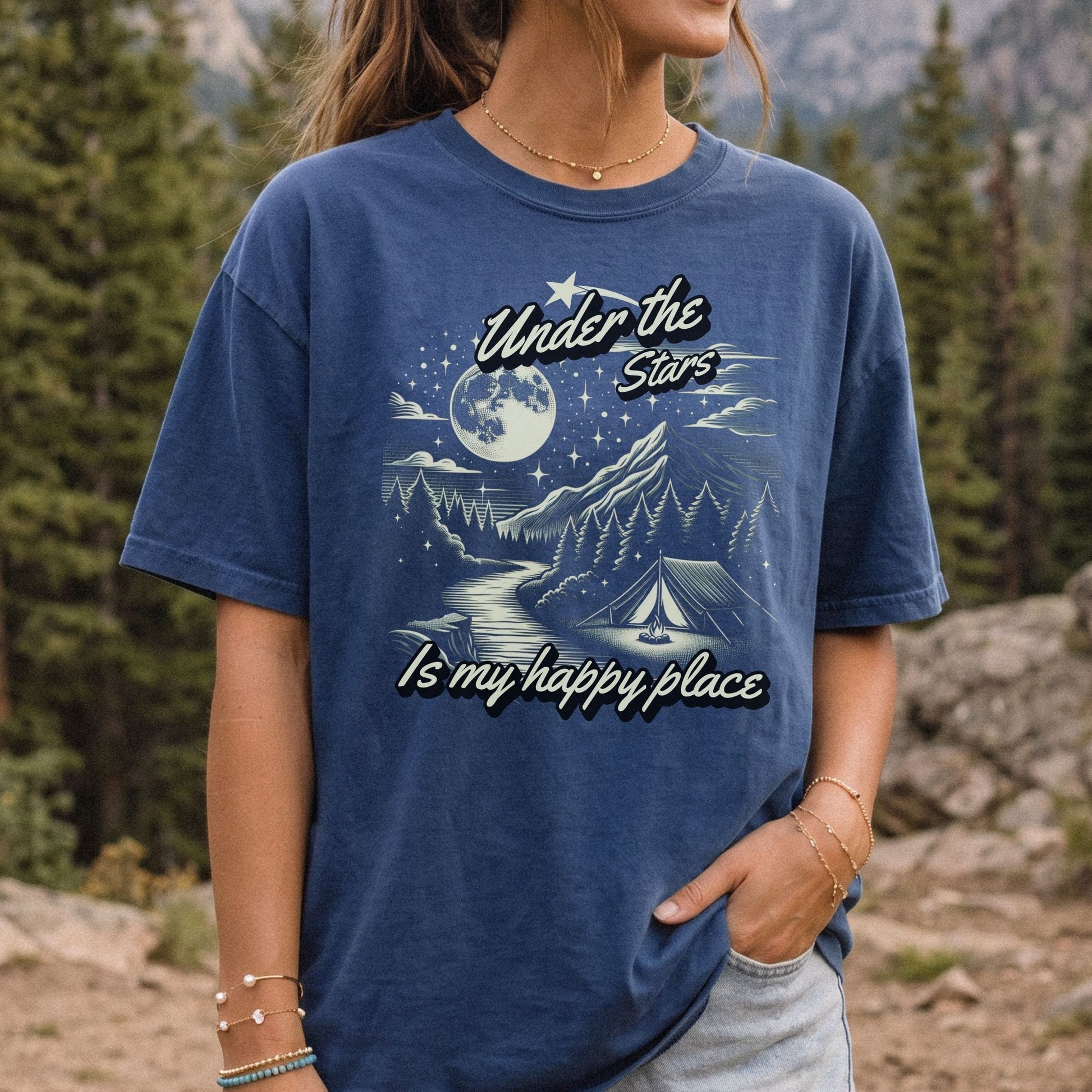A woman wearing a blue graphic t-shirt with a camping moon-and-stars design and the text 'Under the Stars Is my happy place', photographed outdoors in a forest setting.