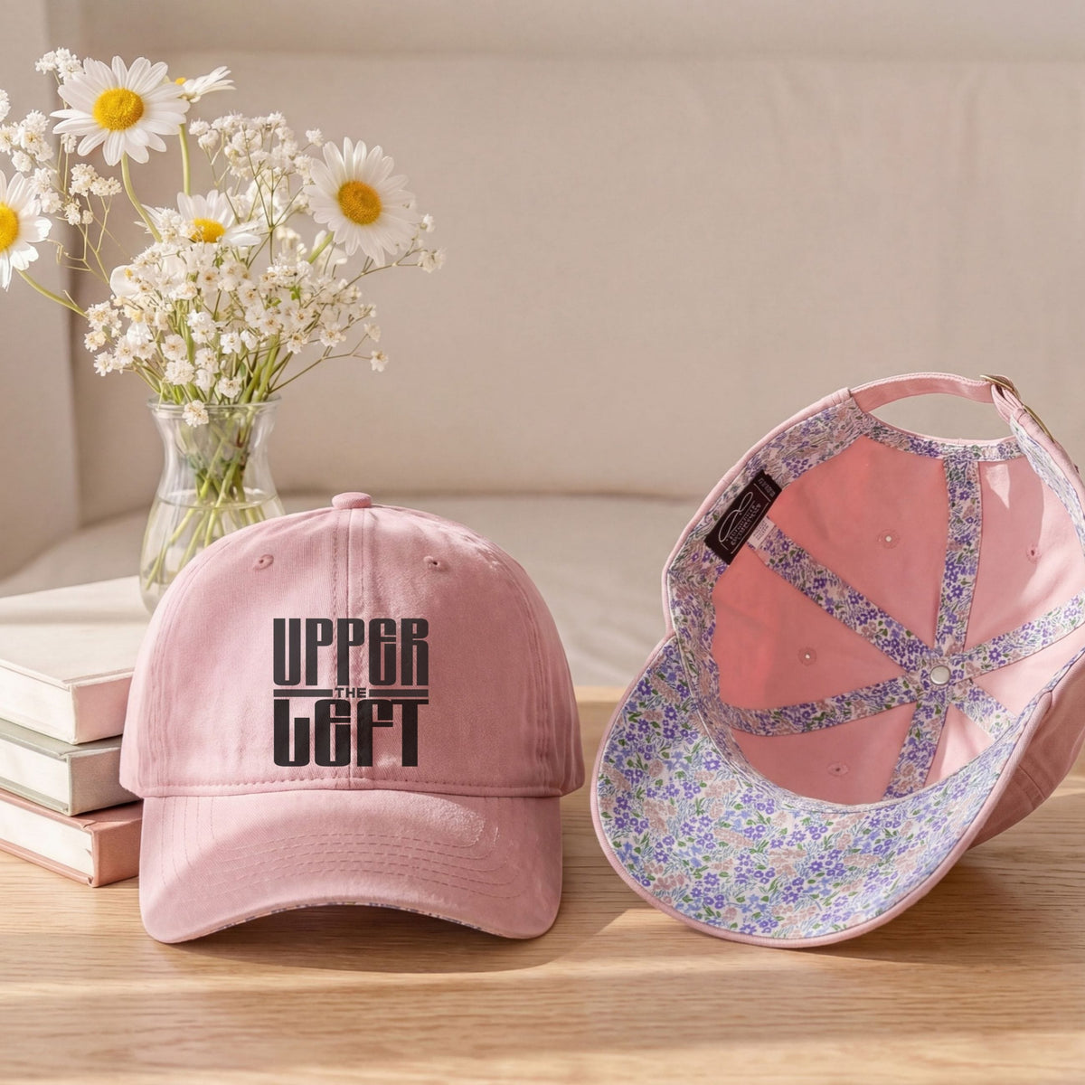 A pink baseball cap shown from the front and inside, styled on a wooden table with a vase of white daisies and stacked books in the background. The front of the cap reads 'The Upper Left' in black text, and the inside features a floral pattern with rhinestone straps.