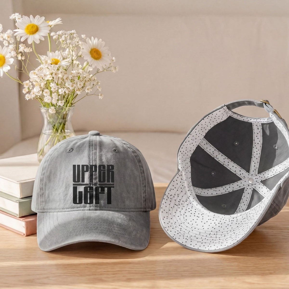 Two gray baseball caps are displayed on a wooden table with a vase of daisies and stacked books in the background. One cap is shown from the front with the text 'UPPER LEFT' printed in black, and the other is shown from the back with a white brim covered in small black dots.