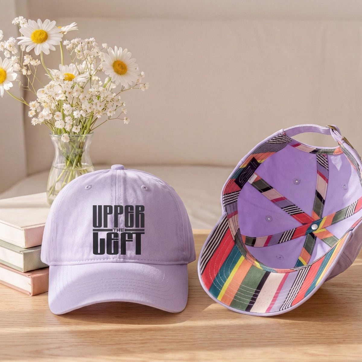 Two baseball caps displayed on a wooden table with a vase of daisies and stacked books in the background. The cap on the left is light lavender with black embroidered text reading 'UPPER LEFT'.