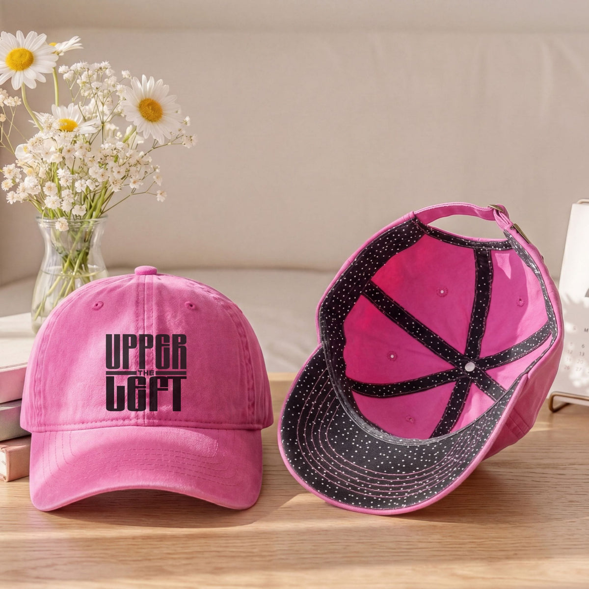 Two pink baseball caps displayed on a wooden table in a styled indoor setting. One cap is shown from the front with black embroidered text reading 'UPPER Left' and the other is shown from the side/back with a black brim and black dotted trim.