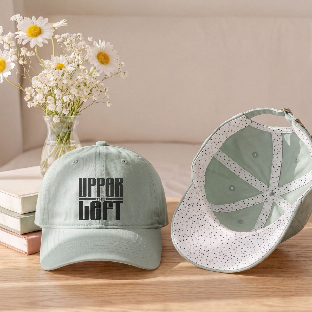 A sage green baseball cap with black text reading 'The UPPER LEFT' sits on a wooden table next to another cap showing its dotted interior. The scene is styled with a vase of white daisies and books in the background.