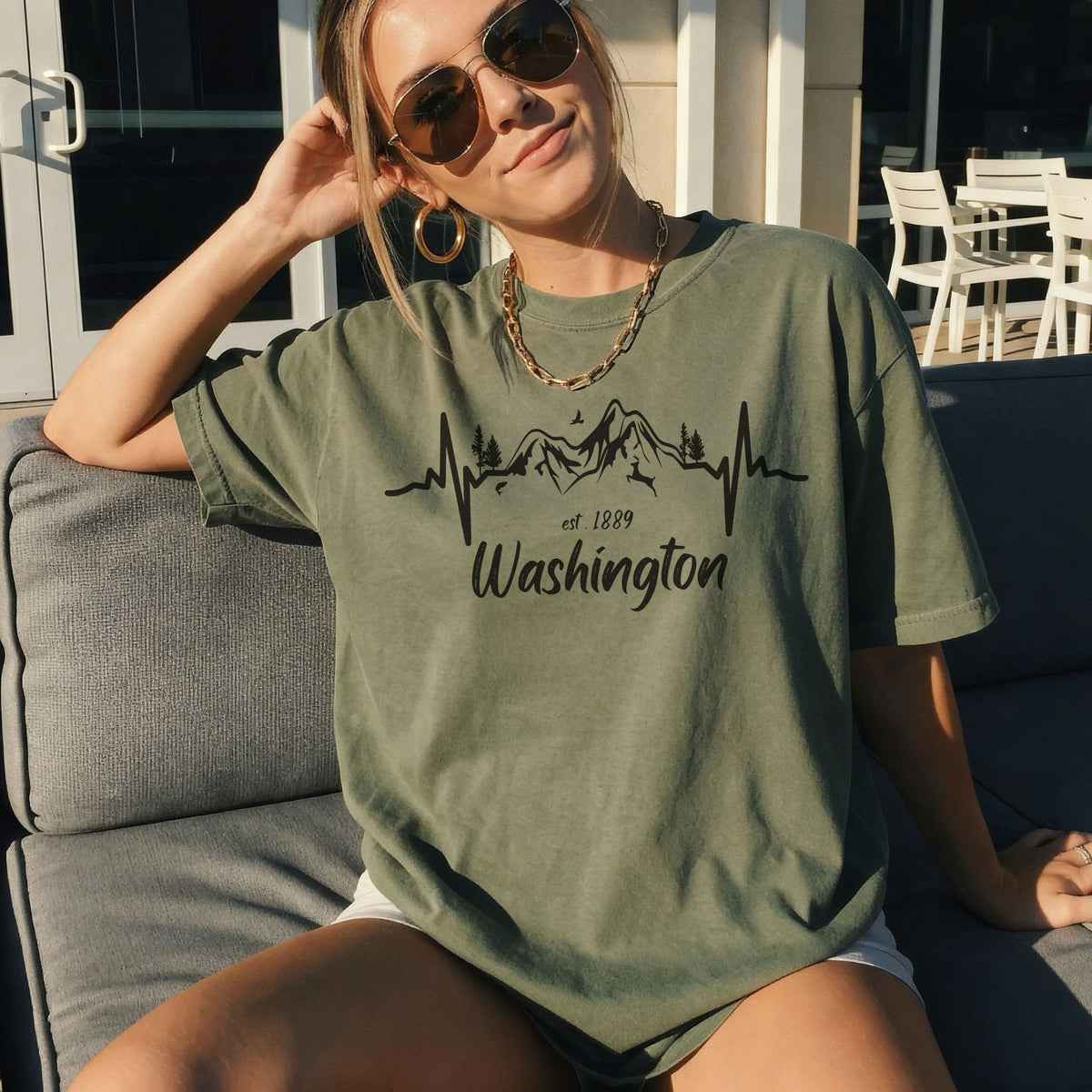 A woman wearing a military green graphic t-shirt with a black mountain and heartbeat line design that reads 'Washington est. 1889', sitting on a gray outdoor sofa.