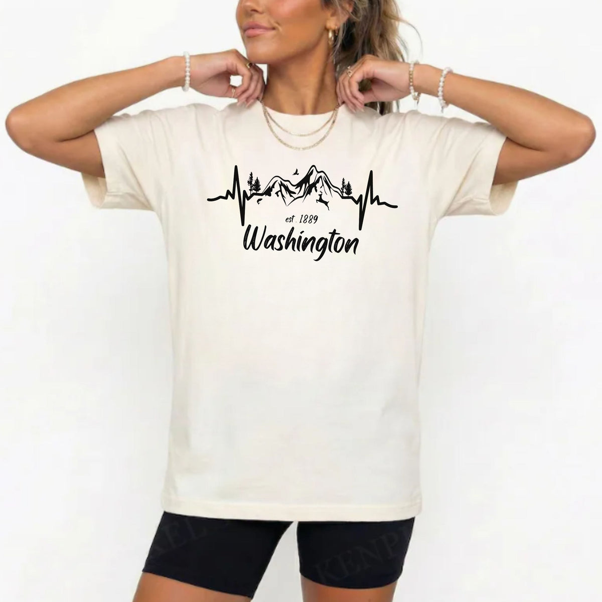 A woman wearing an oversized cream-colored t-shirt with a black graphic of a mountain range on an EKG line and the text 'Washington est. 1889'.