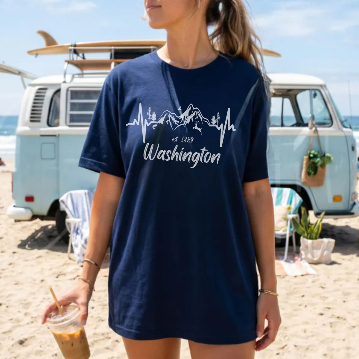 A woman wearing an oversized navy blue t-shirt with a white graphic of a mountain skyline and heartbeat line above the text 'est 1889 Washington', standing on a sandy beach in front of a light blue vintage van with surfboards.