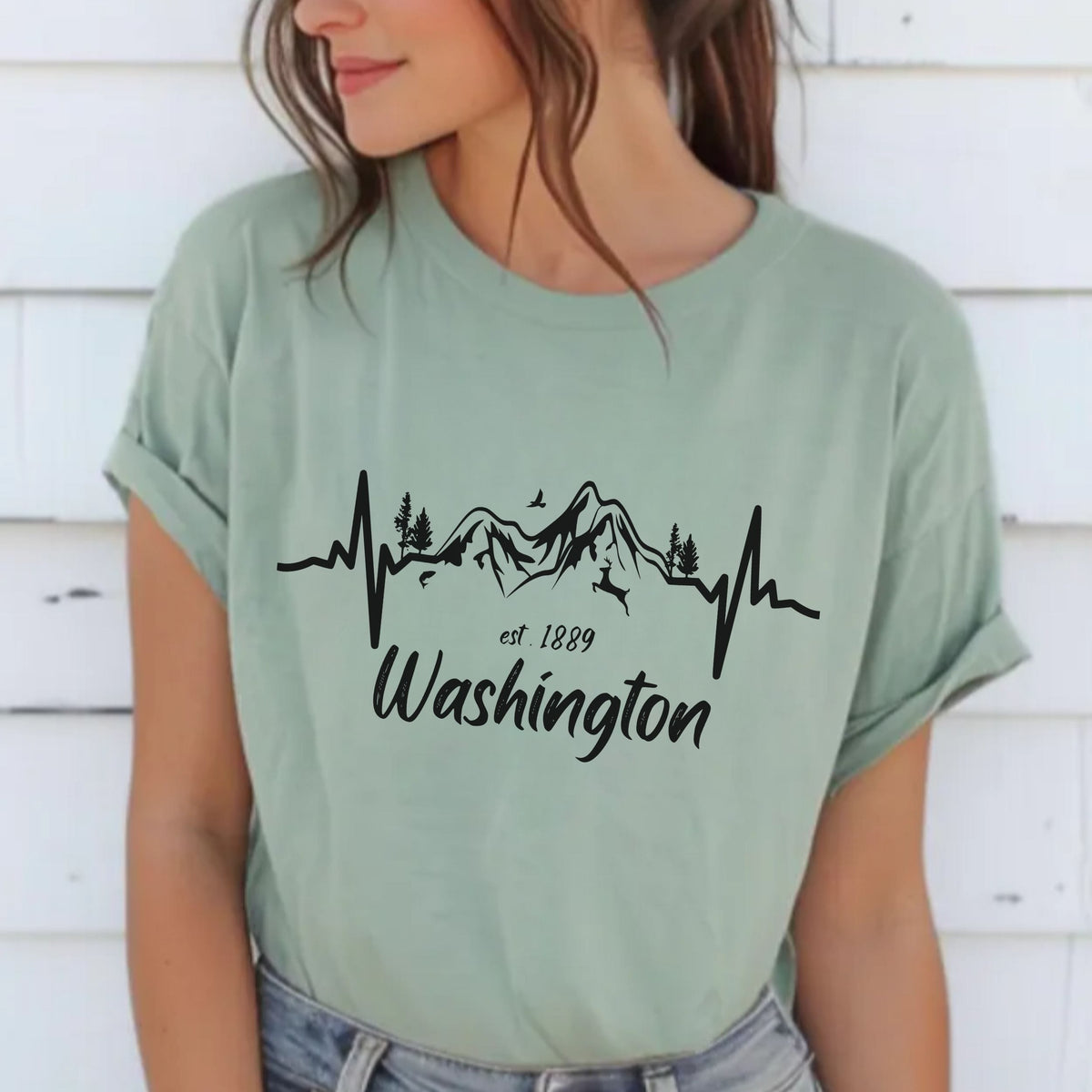 A woman wearing a sage green graphic t-shirt with a black mountain and heartbeat line design that reads 'Washington est. 1889'