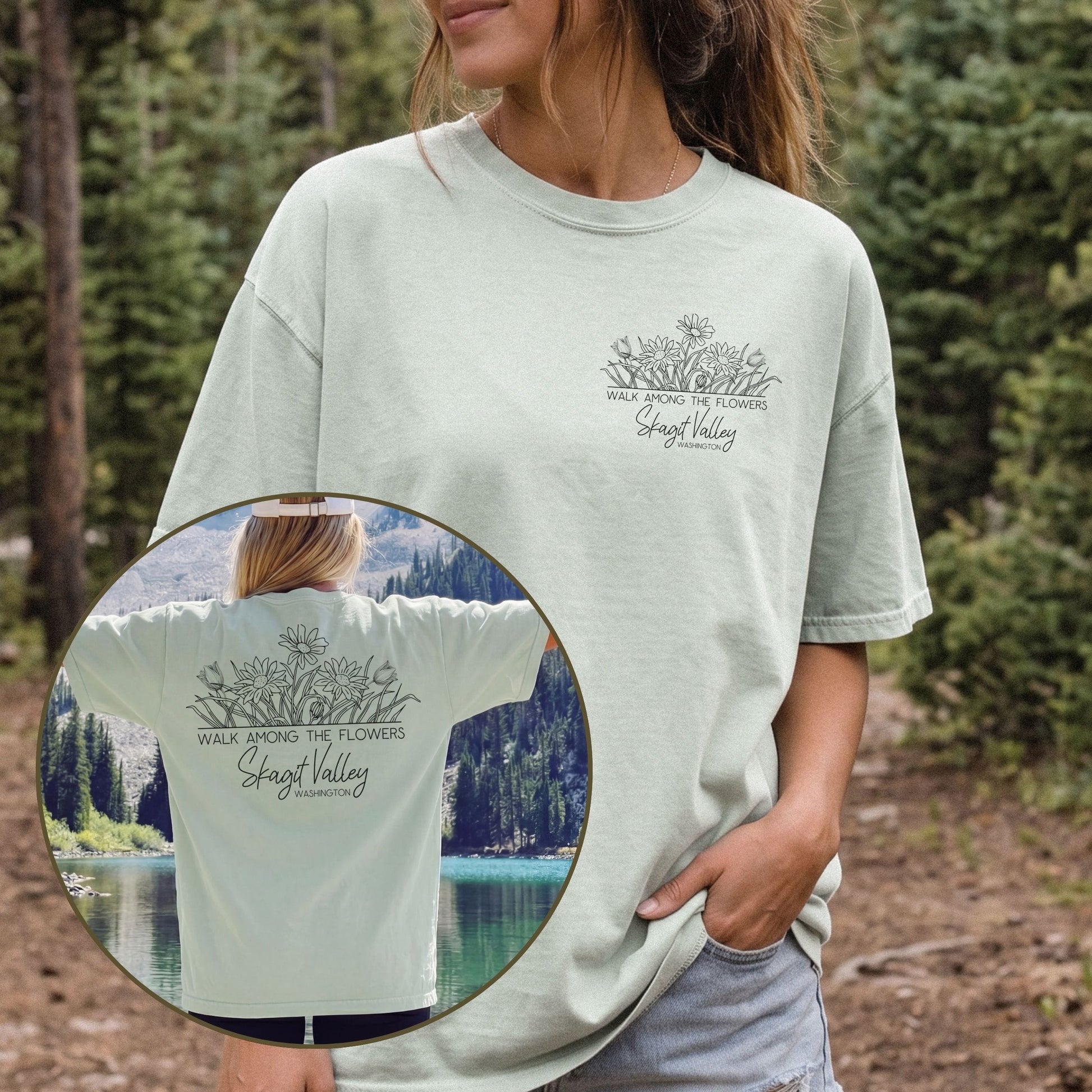 A woman wearing a light sage green graphic t-shirt in an outdoor forest setting, with an inset circle showing the back of the shirt with a larger 'Skagit Valley' graphic. The shirt features a small chest graphic on the front and a larger graphic on the back.