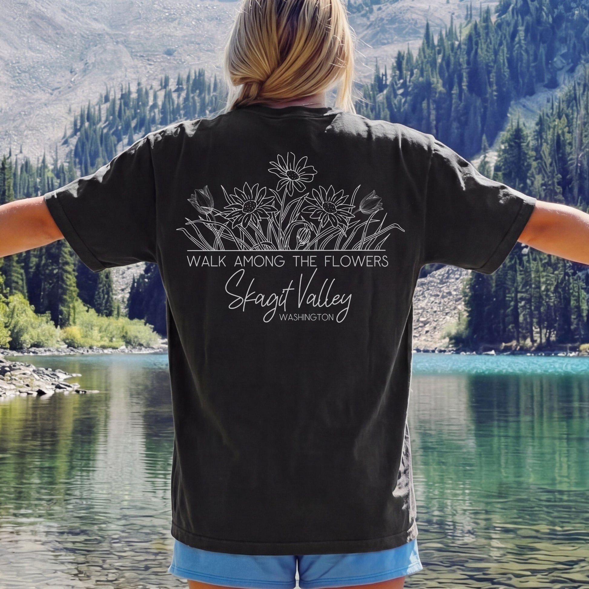 A woman stands with her back to the camera wearing a black graphic t-shirt featuring white floral line art and the text 'WALK AMONG THE FLOWERS', 'Skagit Valley', and 'Washington'. The background shows a scenic mountain lake with pine trees.