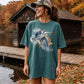 A woman wearing an oversized teal t-shirt with a large heron graphic, standing outdoors near a wooden dock and a rustic cabin in autumn.