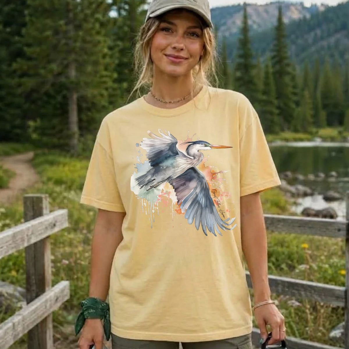 A woman wearing a pale yellow graphic t-shirt with a watercolor heron design, standing outdoors in front of a wooden fence and a forested mountain landscape.