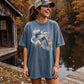 A woman wearing an oversized slate blue graphic t-shirt with a heron print, standing on a wooden dock by a lake with autumn trees in the background.