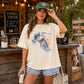 A woman stands at an outdoor rustic campsite or s'mores station wearing a cream-colored graphic t-shirt with a heron print, denim shorts, and a green 'S'mores Station' cap.