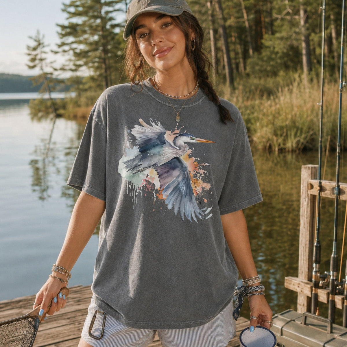A woman stands on a wooden dock by a lake wearing a faded dark gray graphic T-shirt with a large heron print, white shorts, and a gray cap. Fishing rods and a boat are visible in the background.