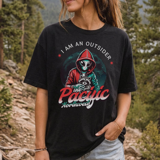 A woman wearing a black graphic t-shirt in an outdoor forest setting. The t-shirt features a graphic of a hooded figure with a mask and the text 'I AM AN OUTSIDER' and 'the Pacific Northwest'.