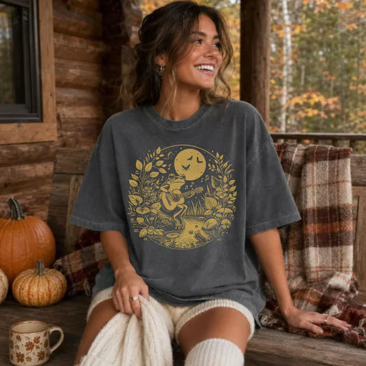 A woman sitting on a rustic wooden porch wearing a dark gray graphic t-shirt with a gold-toned circular illustration of a woman, a cat, and a full moon. The background features a cozy fall theme with pumpkins, a plaid blanket, and a wooden log wall.